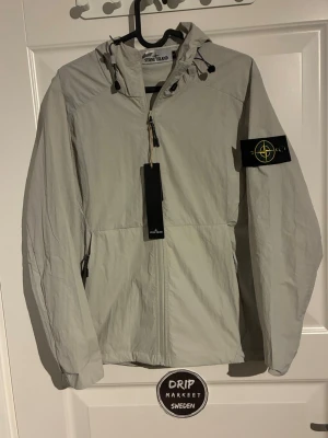 Ljusgrå vindjacka från Stone Island - Snygg ljusgrå vindjacka från Stone Island i storlek small med huva och dragkedja framtill. Jackan har den klassiska Stone Island-loggan på vänster ärm och justerbara snören vid huvan. Tillverkad i lätt och slitstarkt material, perfekt för blåsiga dagar.