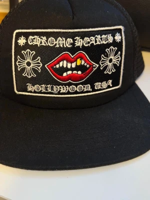 Svart Chrome Hearts keps med patch - Säljer en svart keps från Chrome Hearts med en stor broderad patch framtill. Patchen har motiv av röda läppar med tänder, vita kors och texten 'Chrome Hearts Hollywood, USA'. Klassisk böjd skärm och justerbar baksida.