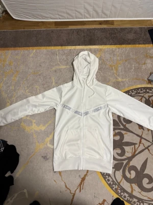 Vit hoodie från Nike med dragkedja - Säljer en vit hoodie från Nike med hel dragkedja och huva. Den har ett grått mönster med Nike-logga över bröstet och två stora fickor framtill. Materialet är mjukt och sportigt, perfekt för en avslappnad look.