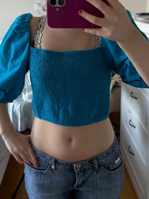 Blå croppad topp med puffärmar - Säljer en blå croppad topp med puffiga kortärmar och smockad framsida. Toppen har en fyrkantig urringning och är perfekt för dig som gillar trendiga och färgglada plagg. Materialet känns lätt och passar bra till jeans eller kjol.