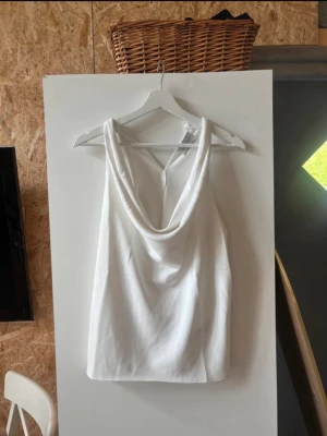 Vit ärmlös linnetopp med drapering - Säljer en stilren vit linnetopp med snygg draperad halsringning. Toppen är ärmlös och har en mjuk, flowy passform som känns både trendig och enkel att matcha. Perfekt för dig som gillar minimalistisk stil.