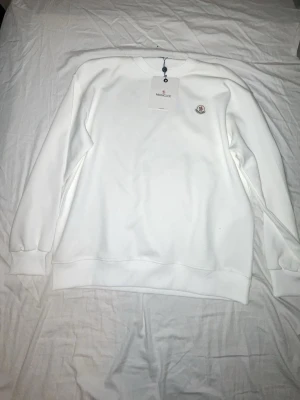Vit sweatshirt från Moncler - Säljer en stilren vit sweatshirt från Moncler med rund halsringning och diskret logga på bröstet. Tröjan har ribbade muddar vid ärmslut och nederkant. Perfekt för dig som gillar klassisk och enkel stil.
