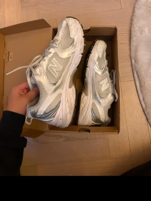 Vita och silver New Balance sneakers - Säljer ett par vita och silverfärgade New Balance sneakers med meshpaneler och reflekterande detaljer. Skorna har en chunky sula och klassisk snörning. Perfekta för dig som gillar sportig och trendig stil.
