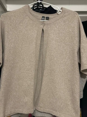 Beige kofta med kort ärm  - Stilren beige kofta  med korta ärmar och rund halsringning. Koftan har en knapp framtill och är tillverkad i ett mjukt, stickat material. Perfekt för lager på lager och enkel att matcha med olika outfits.