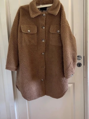 Beige teddyjacka med fickor - Mysig beige teddyjacka med stora bröstfickor och knappar framtill. Jackan har en klassisk krage och är gjord i fluffigt teddy-material som håller dig varm. Perfekt oversized look och längre modell som går ner över höfterna. Snygg detalj med synliga knappar.