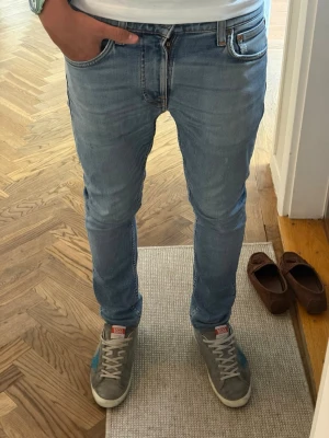 Nudie Lean Dean 28/32 - Säljer ett par ljusblå skinny jeans från Nudie med klassiska fem fickor och snygga kontrastsömmar. Jeansen har en smal passform och är tillverkade i mjukt denimtyg. Perfekta för en avslappnad och trendig look. Jeansen är uppsydda.