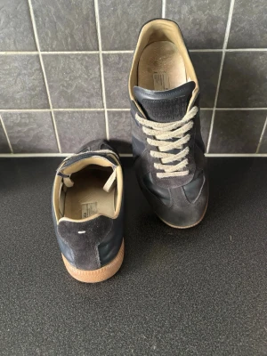 maison margiela skor - Snygga svarta sneakers med detaljer i mocka och släta partier i läder. Skorna har vita snören och en klassisk gummisula i beig. Låg modell med bekväm innersula och stilren design som passar till det mesta.