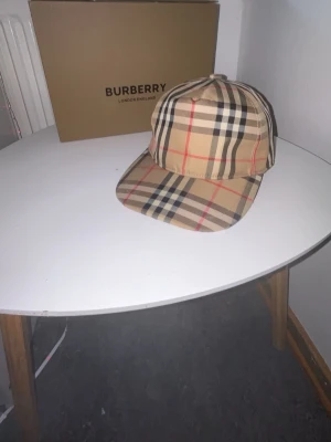 Burberry klassisk rutig keps - Säljer en ikonisk keps från Burberry i deras klassiska beigea rutmönster med röda, svarta och vita detaljer. Kepsen har böjd skärm och justerbar passform. Tillverkad i bomull för en bekväm känsla och snygg look.