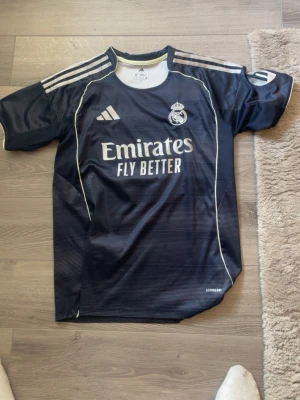  Real Madrid bortamatchtröja - Snygg mörkblå Real Madrid borta matchtröja från Adidas med vita detaljer och klubbmärke på bröstet. Tröjan har vita ränder på axlarna och texten 'Emirates Fly Better' på framsidan. Bellingham på ryggen knappt använd 