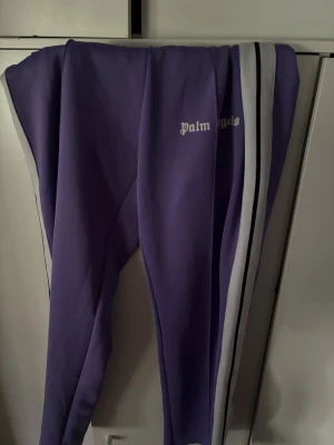 Lila Palm  mjukisbyxor - Säljer ett par lila mjukisbyxor från Palm Angels med vita och svarta ränder längs sidorna. Byxorna har dragkedja vid bensluten och logga tryckt på ena benet. Perfekta för en chill och trendig look.
