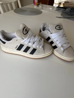 Adidas Campus beige och svarta sneakers - Snygga Adidas Campus sneakers i beige mocka med klassiska svarta ränder på sidorna och vit sula. De är knappt använda någonting så har typ inga defekter förutom det i hälen som syns på bilden😇