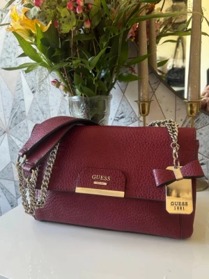 Vinröd väska från Guess - Vinröd väska från Guess i strukturerat konstläder. Väskan har guldfärgade detaljer, kedjerem och en dekorativ rosett med Guess-tag. Oerhört fin väska, jag fick den i gåva men den har tyvärr aldrig kommit till användning. Den är i princip i nyskick, endast en sminkfläck på insidan av fodret från tidigare ägare. Dock går det nog bort med lite sminkborttagning!💞