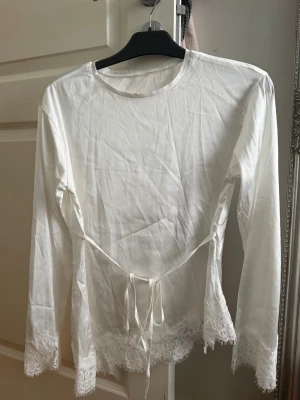 Vit blus med spetsdetaljer och knytband - Snygg vit blus i tunt tyg med långa ärmar. Blusen har fina spetsdetaljer vid ärmslut och nederkant samt ett knytband i midjan för extra stil. Perfekt till jeans eller kjol för en trendig look.