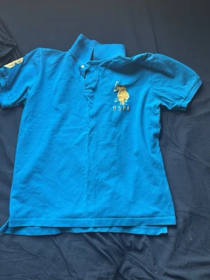 Blå pikétröja med USPA-logga - Snygg blå pikétröja med korta ärmar och klassisk krage. På bröstet finns en broderad gul USPA-logga med polospelare. Tröjan har knappar vid halsen och är perfekt för en avslappnad stil.