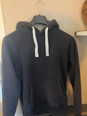 Russemerh mörkblå hoodie - En stilren mörkblå hoodie med vita dragsnören och stor känguruficka framtill. Hoodien har huva och ribbade muddar vid ärmslut och nederkant. Perfekt för en avslappnad look och enkel att matcha med jeans eller joggers. Skriv vid intresse 