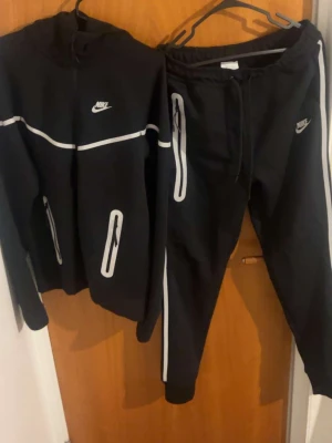 Svart Nike tracksuit med reflexdetaljer - Svart hoodie från Nike med vita reflexdetaljer och dragkedjor. Hoodien har en sportig look med vit Nike-logga på bröstet och reflekterande linjer längs ärmar och fickor. Perfekt för dig som vill sticka ut och synas i mörkret.