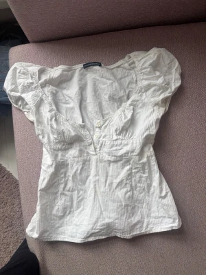 Vit topp med puffärm från Brandy Melville - Söt vit topp från Brandy Melville med puffärmar och djup v-ringning. Toppen har knappar framtill och är gjord i ett tunt, krinklat bomullstyg som känns luftigt och somrigt. Perfekt till jeans eller kjol för en avslappnad look.