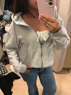 Ljusgrå zip hoodie med huva - Säljer en ljusgrå hoodie med dragkedja och huva. Hoodien har fickor framtill och ribbade muddar vid ärmar och nederkant. Perfekt för en chill och avslappnad stil. Tillverkad i mjukt sweatshirtmaterial.