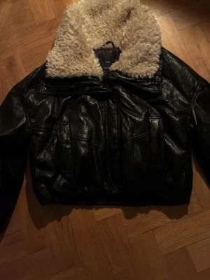 Svart skinnjacka med teddykrage från Mango - Snygg svart skinnjacka från Mango med stor, fluffig teddykrage i beige. Jackan har en kortare modell och flera fickor framtill. Perfekt för dig som vill ha en cool och trendig look med extra värme runt halsen.
