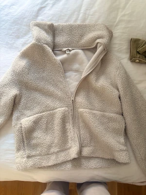Beige teddyjacka med huva, M - Mysig beige teddyjacka med dragkedja framtill och stor huva. Jackan har två stora fickor framtill och är gjord i fluffigt, mjukt material som håller dig varm. Perfekt för kyliga dagar och ger en avslappnad, trendig look.