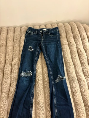 Mörkblå slitna bootcut jeans - Säljer ett par mörkblå bootcut jeans med slitningar och hål på båda benen. Jeansen har vit kontrastsöm och klassisk femficksmodell. Perfekta för en avslappnad och trendig look. Materialet är denim med stretch.jag har gjort hålen själv 