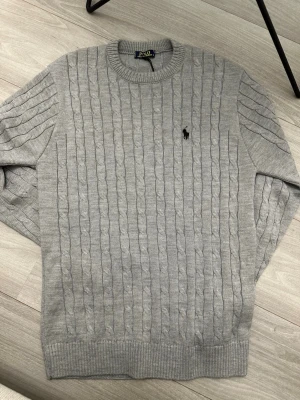Grå kabelstickad tröja Polo Ralph Lauren - Snygg grå kabelstickad tröja från Polo Ralph Lauren med rund halsringning och svart broderad logga på bröstet. Klassisk passform och ribbade muddar vid ärmslut och nederkant. Perfekt till jeans eller chinos.