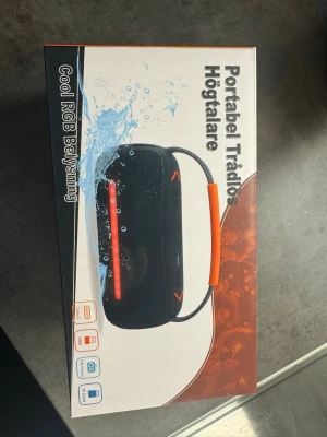 Portable Trådlös Högtalare (Cool RGB Belysning) - This is a portable wireless speaker with cool RGB lighting. It features a smart chip, voice prompts, extra bass, multiple play modes, and universal compatibility. The packaging is in Swedish, indicating it is likely marketed in Sweden or other Scandinavian countries. The speaker is also shown as water-resistant or splash-proof based on the water imagery on the box.