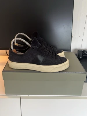 Svarta Tom Ford sneakers i mocka - Snygga svarta sneakers från Tom Ford i mocka med detaljer i läder och vit sula. Klassisk låg modell med svart logga på hälen och svarta skosnören. Perfekta för dig som gillar stilrena och exklusiva sneakers.