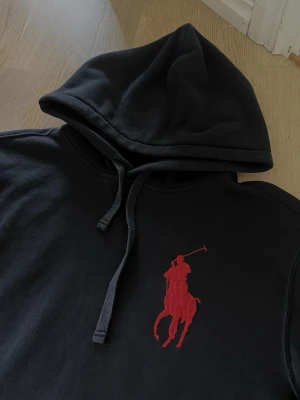 Svart Ralph lauren hoodie - Säljer en svart hoodie från Polo Ralph Lauren med stor röd polospelare broderad på bröstet och en röd siffra på ärmen. Hoodien har huva med dragsko och en klassisk magficka. Perfekt för dig som gillar stilrena plagg.