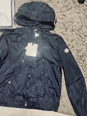  Moncler jacka - Snygg mörkblå vindjacka från Moncler med huva och dragkedja framtill. Jackan har två fickor med tryckknappar och Moncler-logga på ärmen. Tillverkad i glansigt, lätt material och har justerbar dragsko vid huvan.