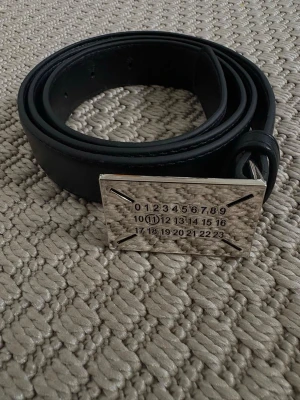Svart bälte med silverfärgat spänne - Stilrent svart bälte i skinn med ett rektangulärt silverfärgat spänne som har en unik numrerad design. Perfekt accessoar för att lyfta din outfit med en modern touch.