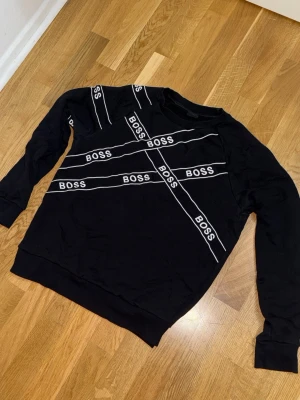 Svart BOSS sweatshirt med logga - Svart sweatshirt från BOSS med vita diagonala loggor över hela framsidan. Tröjan har rund hals, långa ärmar och ribbade muddar. Perfekt för en trendig och avslappnad stil.