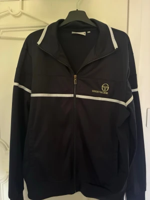Svart träningsjacka Sergio Tacchini - Snygg svart träningsjacka från Sergio Tacchini med vit rand längs bröstet och ärmarna. Jackan har dragkedja framtill, hög krage och logga på vänster bröst. Perfekt till sport eller chill. Tillverkad i ett mjukt och följsamt material.