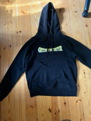 Svart hoodie med tryck Ortens Favoriter - Svart hoodie med stor huva och magficka. På bröstet finns ett gult och grått tryck med texten 'Ortens Favoriter' i streetstil. Hoodien har dragsnören vid huvan och är gjord i mjukt bomullsliknande material. Perfekt för en avslappnad och trendig look. Nästan helt oanvänd! 
