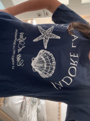 Mörkblå t-shirt med snäckmotiv - Säljer en mörkblå t-shirt från Amisu med vita tryck av en sjöstjärna och snäcka på framsidan samt text. T-shirten har korta ärmar och är gjord i mjuk bomull, perfekt för en chill och avslappnad stil. Den är lång i storleken så förvänta inte dej en magtröja även fast den är S!!!💕💕