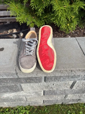 Gråa Louboutin skor  - Snygga gråa sneakers i mocka med klassisk låg profil och vita sulor. Skorna har gråa skosnören och en unik röd undersida som sticker ut. Insidan är beige och materialet är mjukt och bekvämt. Perfekta för dig som vill ha något stilrent och som passar i alla lägen! 