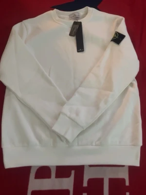 Vit sweatshirt från Stone Island - Säljer en clean vit sweatshirt från Stone Island med den klassiska svarta patchen med gulgrönt broderi på ärmen. Tröjan har rund hals, ribbade muddar och är tillverkad i mjukt bomullstyg. Perfekt för dig som gillar streetwear och vill ha en stilren look.