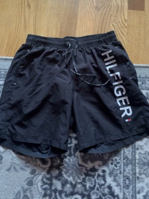 Svarta badbyxor från Tommy Hilfiger - Snygga svarta badbyxor från Tommy Hilfiger med stort vitt HILFIGER-tryck längs benet och liten logga i rött, vitt och blått. Resårmidja med dragsko och sidofickor. Perfekta för stranden eller poolen.