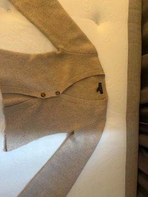 Beige kofta från Amisu med knappar - Mysig beige kofta från Amisu med två träknappar framtill. Koftan har v-ringning och är stickad i ett mjukt material som passar perfekt till lager på lager. Enkel och stilren design som är lätt att matcha.