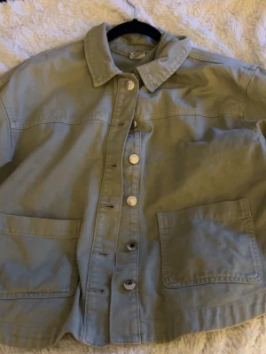 Ljusgrön jeansjacka från Vero Moda - Trendig ljusgrön jeansjacka från Vero Moda med klassisk krage, stora framfickor och silverfärgade knappar. Jackan har en avslappnad passform och är tillverkad i tåligt denimtyg. Perfekt för att lyfta din outfit med en cool touch.