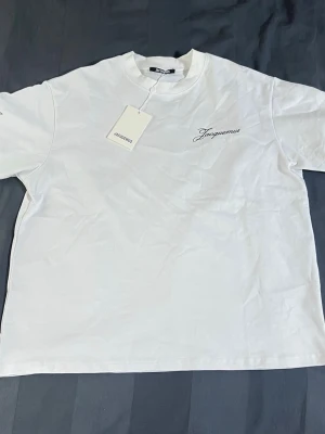 Vit Jacquemus t-shirt i bomull - Snygg vit t-shirt från Jacquemus med svart broderad logga på bröstet. Klassisk rund hals och korta ärmar. Tillverkad i mjuk bomull som känns skön mot huden. Perfekt till jeans eller shorts för en clean och stilren look.