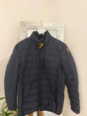Mörkblå pufferjacka från Parajumpers - Snygg mörkblå pufferjacka från Parajumpers med gul detalj vid kragen och klassisk patch på ärmen. Jackan har dragkedja framtill, två sidofickor med dragkedja och en bröstficka. Perfekt för kalla dagar och riktigt skön passform. Priset kan sänkas vid snabba äffärer 