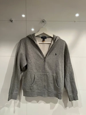 Ralph Lauren zip hoodie - Hej, säljer en grå Ralph Lauren zip hoodie i storlek L (14-16) men passar även XS. Skick 9/10 använd fåtal gånger. Har inga defekter eller luktar inte konstigt. Vid fler frågor eller funderingar tveka inte att höra av dig. Priset kan diskuteras vid snabb affär!