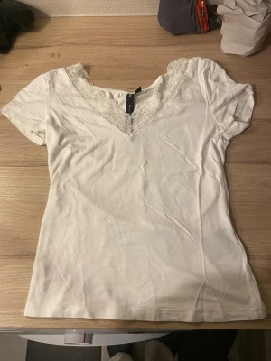 Vit topp med spetsdetalj - Säljer en vit topp med v-ringning och fin spetskant runt halsen. T-shirten har korta ärmar och är gjord i mjukt bomullsmaterial. Enkel och stilren modell som passar till mycket.