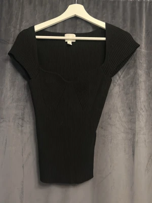 Svart ribbad topp med fyrkantig ringning - Snygg svart ribbad topp med fyrkantig ringning och korta ärmar. Toppen har en figurnära passform och markerade sömmar framtill som ger en modern look. Perfekt att styla med jeans eller kjol för en trendig outfit.