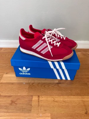 Röda Adidas sneakers med vita ränder - Snygga röda Adidas sneakers med klassiska vita ränder på sidorna och vita skosnören. Skorna har en retroinspirerad design med låg profil och yttersula i gummi. Ovandelen är i textil och mocka för en cool och sportig look.