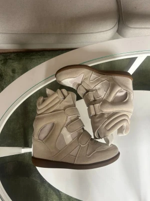 Isabel Marant - Unika beige sneakers med hög siluett och flera breda kardborreband. Skorna har en chunky design med paneler i mocka och syntet, samt en brun sula. Perfekta för dig som vill sticka ut med en cool och modern look.