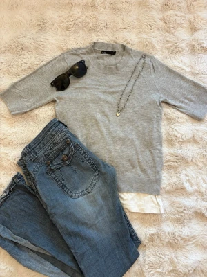 Ljusgrå stickad tröja 157 XS - Ljusgrå stickad tröja från 157 med korta ärmar och ribbstickad halsringning. Tröjan är enkel och stilren, perfekt att matcha med jeans eller kjol. Materialet är mjukt och behagligt, och passar dig som gillar en clean look. Obs! Endast tröjan som tillkommer i denna annons. Jeansen sälj separat.