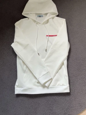 Vit hoodie Prada  - Snygg vit hoodie från Prada med klassisk huva och dragsko. På bröstet finns en röd logga med texten PRADA. Hoodien har en känguruficka framtill och stilrena detaljer i metall på snörena. Perfekt för en clean och trendig look. Aldrig använd splittrans ny