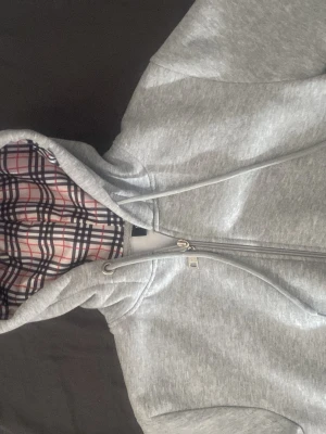 Burbery hoodie - Snygg grå zip-hoodie med dragkedja och huva. Insidan av huvan har ett klassiskt rutigt mönster i svart, vitt och rött. Hoodien har dragsko i huvan och är tillverkad i mjukt sweatshirtmaterial. Perfekt för en chill och avslappnad stil.